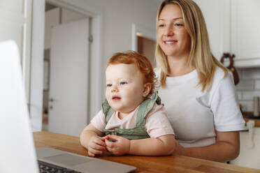 Woman with baby daughter using laptop at kitchen table - VYF00403