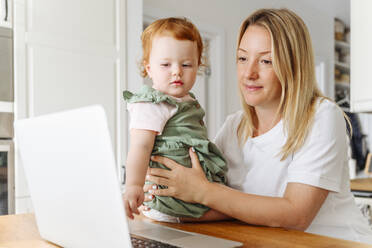 Woman with baby daughter using laptop at kitchen table - VYF00404