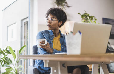 Woman holding coffee cup, sitting in front of laptop at home - UUF22454