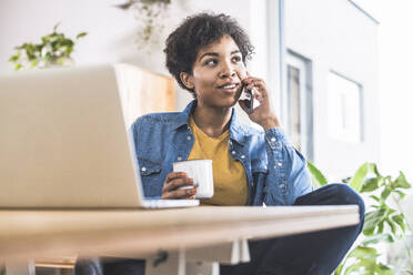 Woman talking on mobile phone, sitting in front of laptop at home - UUF22457