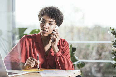 Woman talking on mobile phone and looking at laptop at desk - UUF22462