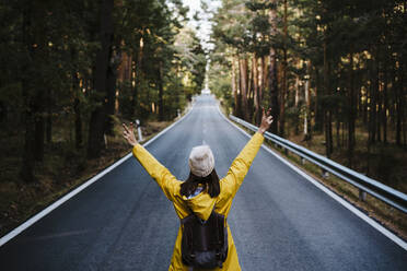 Woman in yellow raincoat standing on empty treelined road - EBBF02097