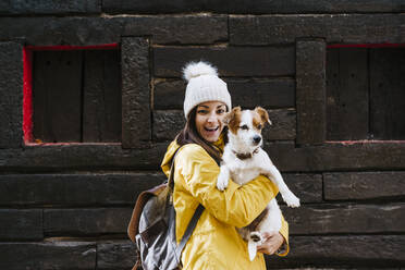 Portrait of smiling woman in yellow raincoat holding dog in front of log cabin - EBBF02098