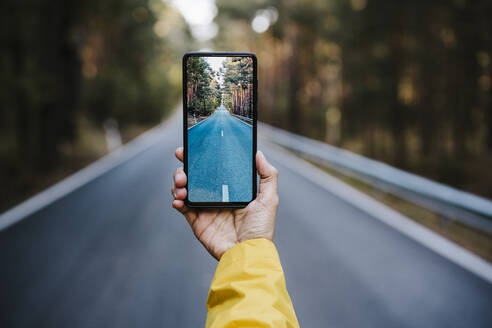 Womans hand holding smart phone, photographing empty country road - EBBF02107