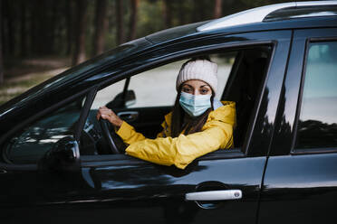 Woman wearing yellow raincoat and face mask sitting in car - EBBF02117