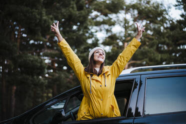 Woman wearing yellow raincoat standing in car with arms raised - EBBF02118