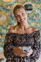 Porträt einer erwachsenen Frau, die mit einem Smartphone in der Hand in die Kamera lächelt - DLTSF01475
