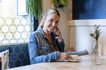 Porträt einer erwachsenen Frau, die in die Kamera lächelt, während sie an einem Restauranttisch sitzt und mit einem Smartphone telefoniert - DLTSF01480
