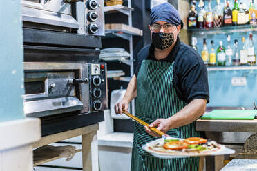 Porträt eines männlichen Kochs mit Schutzmaske, der eine Schaufel mit frisch gebackener Pizza hält - DLTSF01485