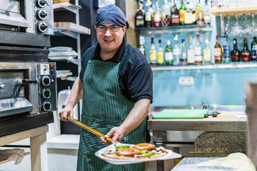 Porträt eines männlichen Kochs, der eine Schale mit frisch gebackener Pizza hält - DLTSF01486