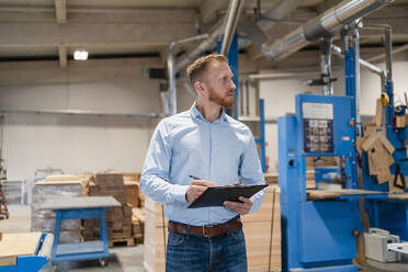 Portrait of carpenter standing in production hall with clipboard in hands - DIGF14289