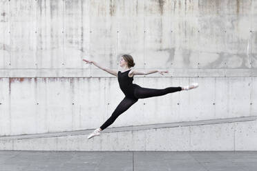 Ballerina in black leotard jumping in front of concrete wall - JPTF00643