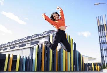 Portrait of beautiful brunette jumping in front of colorful building - JCCMF00752