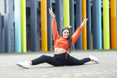 Portrait of beautiful brunette doing splits in front of colorful building - JCCMF00783