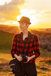 Vorderansicht einer androgynen weiblichen Reisenden mit professioneller Fotokamera, die auf einem Hügel in einer bergigen Gegend steht - ADSF19729