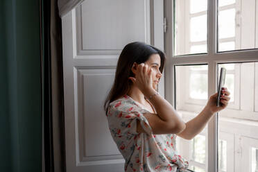 Seitenansicht einer ruhigen Frau mit Smartphone, die am Fenster steht und die Straße durch das Glas fotografiert - ADSF19732