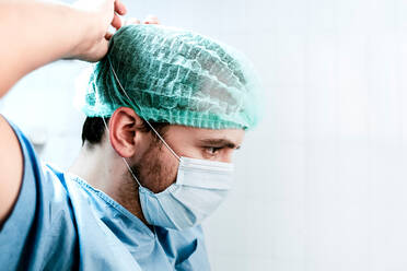 Seitenansicht eines seriösen männlichen Chirurgen in Uniform und Mütze, der eine medizinische Maske aufsetzt, während er sich auf eine Operation im Krankenhaus vorbereitet - ADSF19737