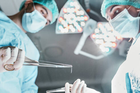 From below side view of focused professional surgeon and nurse with medical scissors working together in operating room in modern hospital - ADSF19738