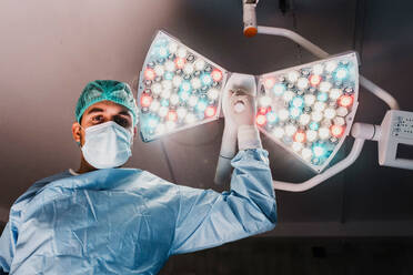 Von unten ernster männlicher Chirurg in Schutzmaske und medizinischer Uniform, der eine chirurgische Lampe einstellt, während er sich auf eine Operation in einem modernen Operationssaal vorbereitet - ADSF19739