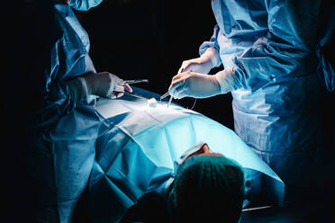 Anonymer Chirurg und Assistent mit chirurgischem Werkzeug und Faden, der die Wunde eines Patienten während einer Operation im Krankenhaus näht - ADSF19742
