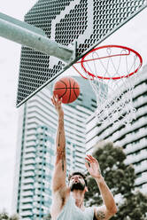 Entschlossener junger männlicher Basketballspieler in Sportkleidung springt und schießt den Ball in der Nähe des Reifens, während er allein auf einem Spielplatz auf der Straße trainiert - ADSF19744