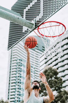 Entschlossener junger männlicher Basketballspieler in Sportkleidung springt und schießt den Ball in der Nähe des Reifens, während er allein auf einem Spielplatz auf der Straße trainiert - ADSF19744