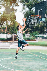 Full body side view of determined young male basketball player in sportswear leaping and shooting ball near hoop while training alone on street playground - ADSF19745