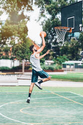 Ganzkörper-Seitenansicht eines entschlossenen jungen männlichen Basketballspielers in Sportkleidung, der springt und den Ball in der Nähe des Reifens schießt, während er allein auf einem Spielplatz auf der Straße trainiert - ADSF19745