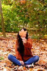 Glückliche junge Brünette in Strickpulli und Jeans versteckt Gesicht hinter gefallenem Ahornblatt, während sie auf buntem Laub im Herbstpark sitzt - ADSF19753