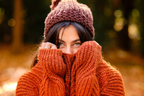 Young female in knitted hat covering face with warm sweater while standing in forest in cold autumn day stock photo