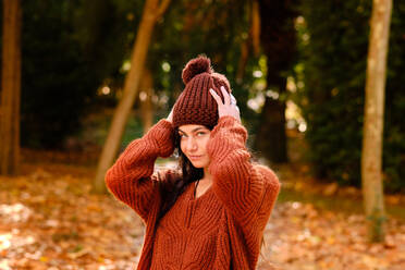 Junge Frau mit Strickmütze und warmem Pullover, die im Wald an einem kalten Herbsttag steht und in die Kamera schaut - ADSF19755