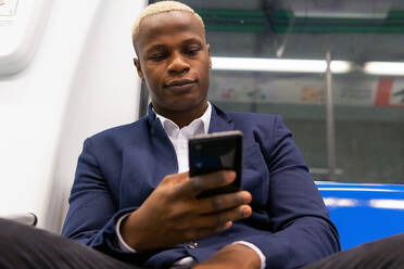 Von unten Afrikanischer männlicher Unternehmer im Anzug, der in einem modernen U-Bahn-Wagen sitzt und auf dem Weg zur Arbeit mit seinem Handy telefoniert - ADSF19787