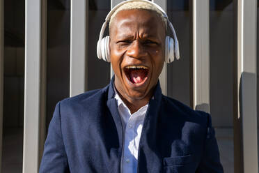 Verträumter afroamerikanischer männlicher Unternehmer im Anzug, der in der Stadt steht und Lieder über kabellose Kopfhörer genießt - ADSF19788