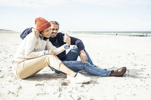 Junges Paar sitzt zusammen am Strand und benutzt ein Smartphone - UUF22524
