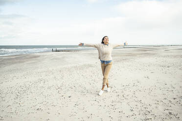 Portrait of young woman walking on sandy beach with raised arms - UUF22534