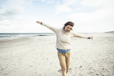 Porträt einer jungen Frau, die mit erhobenen Armen am Sandstrand spazieren geht - UUF22535