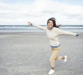 Portrait of young woman running on sandy beach with raised arms - UUF22537