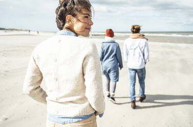 Portrait of young woman walking on sandy beach with two people in background - UUF22541