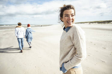 Portrait of young woman walking on sandy beach with two people in background - UUF22542