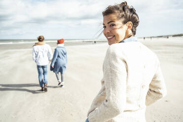 Porträt einer jungen Frau, die am Sandstrand spazieren geht, mit zwei Personen im Hintergrund - UUF22543