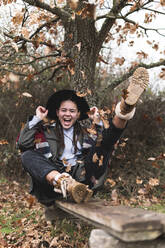 Lächelndes Teenager-Mädchen mit Hut, das auf einer Planke in einer Herbstlandschaft sitzt - JAQF00175