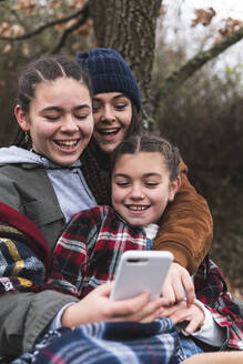 Drei lächelnde Schwestern schauen auf ihr Smartphone in einer Herbstlandschaft - JAQF00181