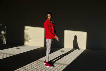 Junger Mann in roter Jacke vor einem Betongebäude im Sonnenlicht - AODF00208
