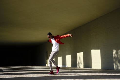 Young man in red jacket in front of concrete building in sunlight - AODF00217