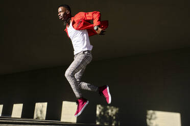 Junger Mann in roter Jacke springt vor einem Betongebäude im Sonnenlicht - AODF00218