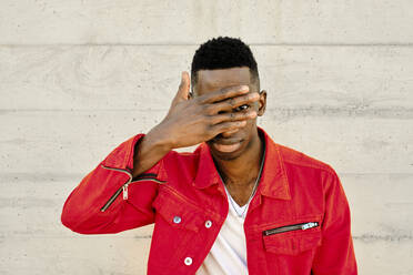 Young man in red jacket covering eyes against concrete wall - AODF00224