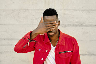 Young man in red jacket covering eyes against concrete wall - AODF00225