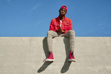 Porträt eines jungen Mannes in roter Jacke und Baseballmütze an einer Betonwand sitzend - AODF00236