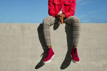 Mann in roter Jacke, karierter Hose und roten Schuhen sitzt an einer Betonwand - AODF00237