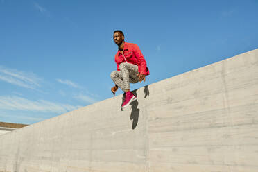 Young man in red jacket and checked pants jumping from concrete wall - AODF00241